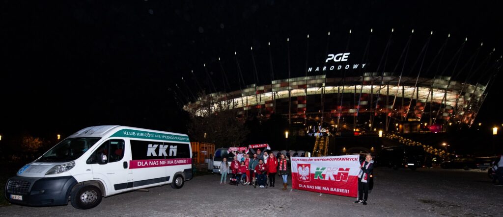 Zmierzch na dworze. Grupa kibiców niepełnosprawnych ubrana w biało-czerwone barwy stoi przed oświetlonym Stadionem Narodowym. Obok nich bus.